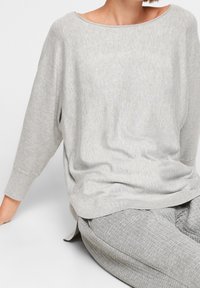 Šedý pletený top s širokým lodním výstřihem, dlouhými rukávy a uvolněným střihem. Látka má jemnou strukturu, kombinovaná s texturovanými šedými kalhotami.