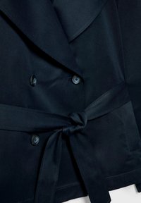 Gros plan d'un manteau bleu marine foncé avec des boutons noirs et une ceinture en tissu nouée à la taille, montrant les revers et une partie d'une poche.