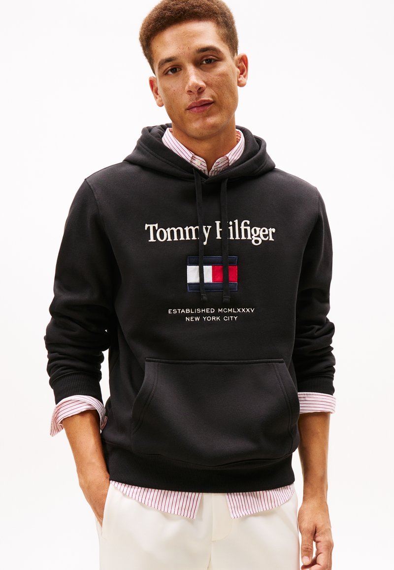 Felpa nera con cappuccio con scritta bianca "Tommy Hilfiger", caratterizzata da un logo ricamato in rosso, bianco e blu. Include una tasca frontale e coulisse.