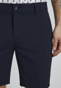 Námořnické modré šortky vyrobené z hladkého materiálu, s předním knoflíkovým zapínáním, plochým předním designem a bočními kapsami. Krátká délka, přiléhavý střih.
