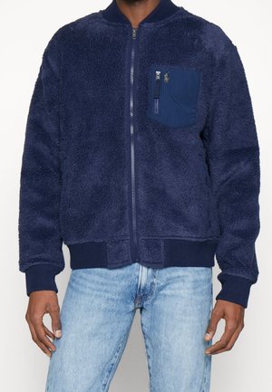 Homme portant une veste polaire bleu marine zippée avec poignets et bas côtelés, dotée d'une poche poitrine zippée, associée à un jean bleu clair.