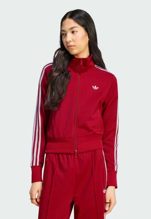 Frau mit langen dunklen Haaren, die einen roten Adidas-Trainingsanzug mit weißen Streifen trägt, steht vor einem einfarbigen hellen Hintergrund.