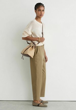 Kvinde iført beige strikket sweater, khakibukser, mønstrede flade sko, bærende en beige spandtaske med brun rem på skulderen foran en hvid væg.