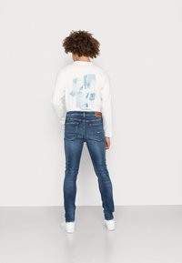 Camisa blanca de manga larga con un gráfico azul en la espalda, combinada con jeans azul oscuro ajustados y zapatillas blancas.