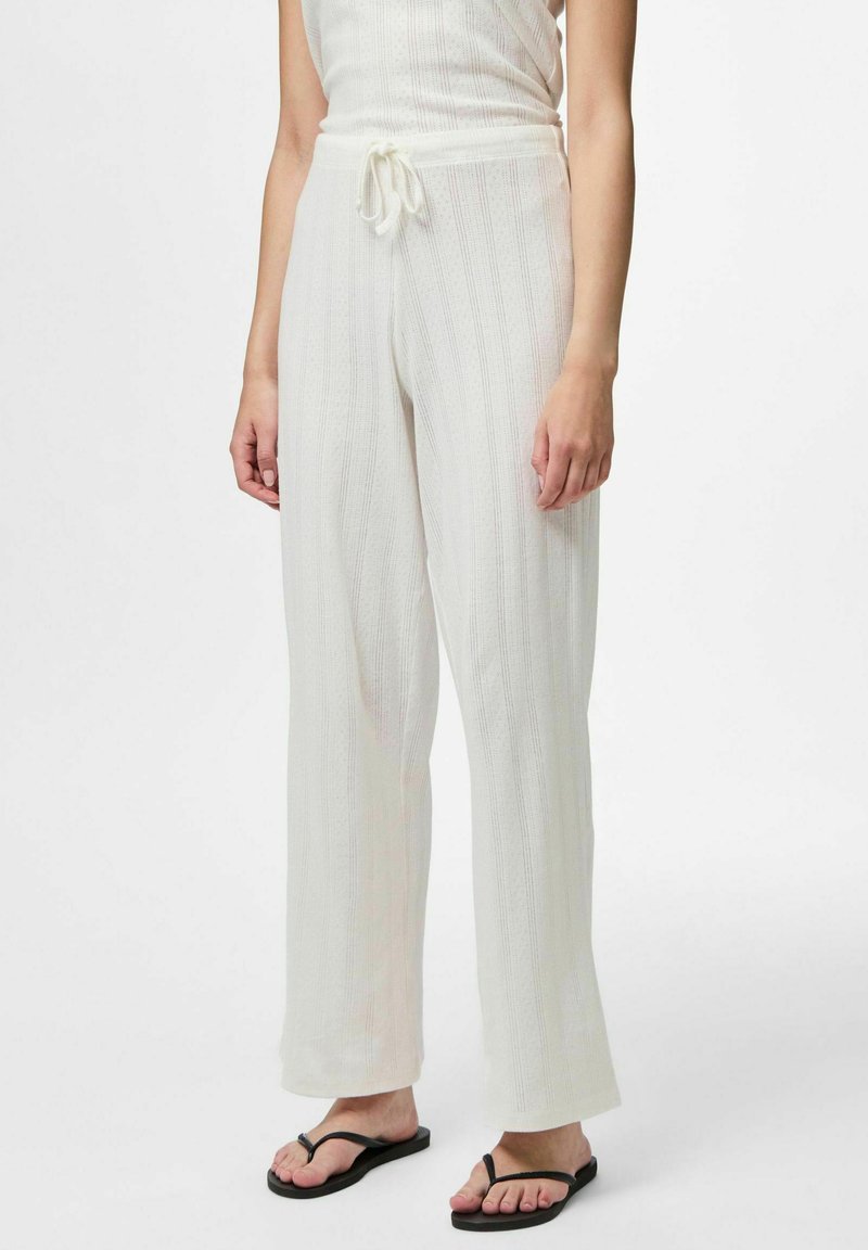 Pantaloni bianchi leggeri e strutturati con una vestibilità ampia, vita con coulisse e righe verticali, abbinati a ciabatte nere.