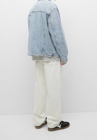 Personne portant une veste en denim bleu clair, un pantalon large blanc et des baskets noires et blanches, debout contre un mur blanc.