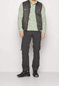 Gilet isolato nero con tasche frontali sopra una camicia a maniche lunghe a motivo verde chiaro, abbinato a pantaloni cargo grigi e scarpe da ginnastica nere.