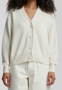 Kobieta w beżowym kardiganie zapinanym na guziki z długimi rękawami oraz pasujących beżowych spodniach, stojąca na tle jednolitego szarego tła.