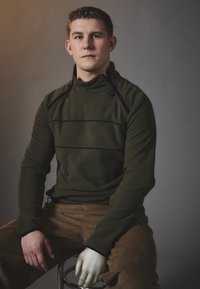 Olivově zelený fleecový pulover s vysokým l�ímcem, černými akcenty a horizontálními švy, kombinovaný s hnědými kordovým kalhotami. Hladká textura.