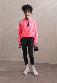 Jeune femme aux cheveux bouclés portant une veste rose vif, des leggings noirs, des chaussettes blanches et des baskets, tenant un masque de fitness à la main.