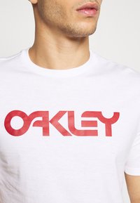 Bílá bavlněná tričko s crew výstřihem, na které se nachází velké červené logo "OAKLEY" v tučném, moderním písmu. Hladká textura.