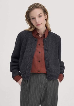 Donkergrijze gebreide cardigan met een ronde hals, geribbelde manchetten en knoopsluiting, gelaagd over een roestkleurige overhemd met knopen.