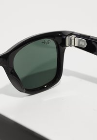 Occhiali da sole neri con lenti verde scuro. La montatura in plastica spessa presenta una cerniera in argento con un logo accentato. Texture liscia e design contemporaneo.