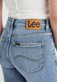 Κοντινό πλάνο ατόμου που φοράει ανοιχτό μπλε τζιν Lee και λευκή ριγωτή μπλούζα, με έμφαση στην πίσω τσέπη και την δερμάτινη ετικέτα της μάρκας Lee στη ζώνη.