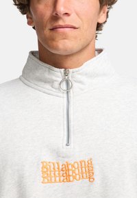 Jasnoszary polarowy pulower z wysokim kołnierzem i półzamkiem. Ma pomarańczowe haftowane logo "Billabong" na piersi. Miękka tekstura.
