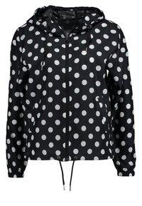 Černá větrovka s velkými bílými puntíky, nastavitelnou kapucí, předním zipem a elastickými manžetami. Obsahuje šňůrku na dolním lemu.