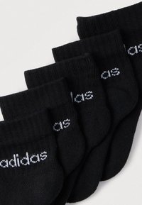 Cinco pares de calcetines negros de tobillo con el logo blanco de Adidas, dispuestos superpuestos sobre una superficie clara.