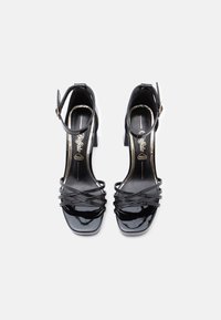 Fekete lakkbőr magas sarkú szandál állítható bokapánttal, négyzet alakú orral és keresztezett pántos kialakítással. Arany színű díszítés a talpbetéten.