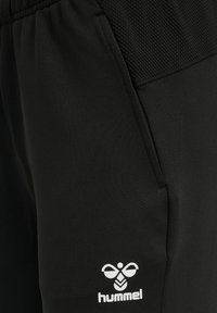 Czarne spodnie sportowe z elastycznym paskiem w talii, teksturowanym górnym panelem, boczną kieszenią i białym logo pszczoły Hummel obok kieszeni.