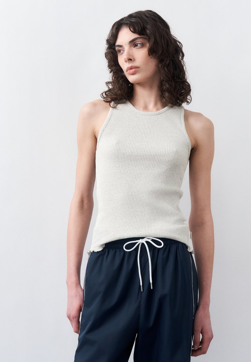 Νεαρή γυναίκα με σγουρά μαλλιά που φοράει ένα μπεζ αμάνικο ριμπ τοπ και μπλε παντελόνι με κορδόνι, κοιτάζοντας πλάγια σε ένα απλό φόντο.