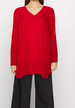 Femme portant une tunique rouge à manches longues ample avec un col en V, associée à un pantalon noir à jambes larges, sur un fond blanc.