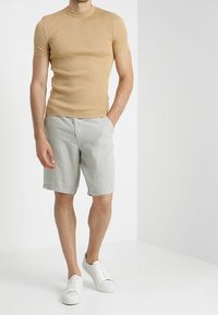 Beżowa, prążkowana koszulka z krótkim rękawem w połączeniu z jasnoszarymi szortami i białymi trampkami. Model stoi w neutralnym otoczeniu na jednolitym tle.