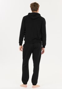 Schwarzer Hoodie und passendes Jogger-Set aus weichem Stoff, mit Kapuze und elastischen Bündchen, von hinten mit baren Füßen gesehen.