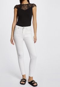 Top negro sin mangas con detalle de encaje en los hombros, combinado con jeans skinny blancos y sandalias negras de deslizar.