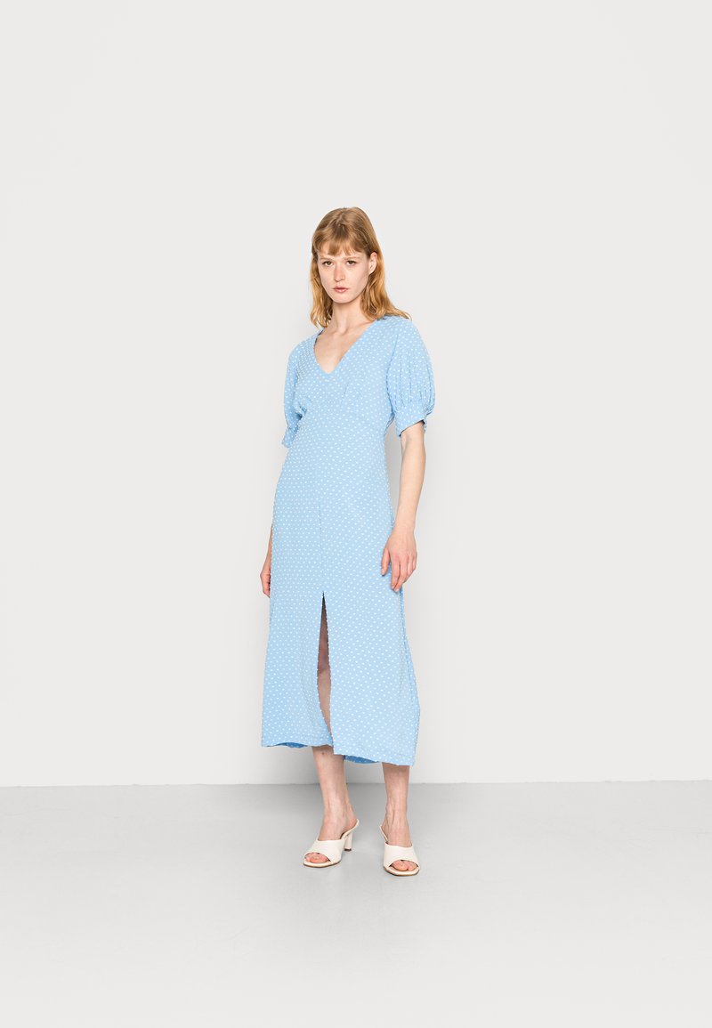 Vestido azul-claro com mangas curtas bufantes, padrão de bolinhas, decote em V e racha frontal, combinado com mules brancos de bico aberto.