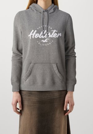 Gray hoodie with a front pocket, featuring white "Hollister" logo, adjustable drawstring hood, and ribbed cuffs. Smooth fabric texture.