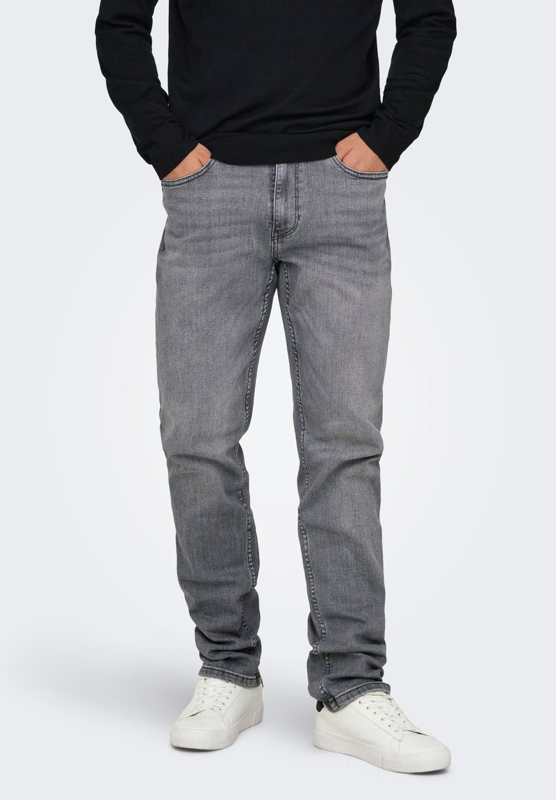 Jean en denim gris à coupe slim, avec cinq poches et des détails de couture subtils, associé à des baskets blanches, porté avec un pull noir.