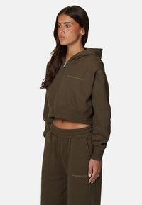 Sweat à capuche vert olive avec fermeture éclair à l'avant, capuche et logo discret. Associé à un pantalon de survêtement assorti avec un waistband élastique.