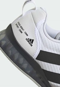 Bijela atletska cipela s teksturiranim gornjim dijelom, crnim trakama i prozirnom potplatom. Značajke uključuju otisnute detalje "VISINA ULOŽKA" i "ODSTUPANJE".