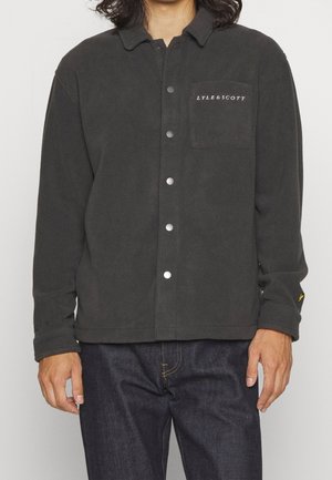 Mężczyzna w czarnej polarowej kurtce zapinanej na guziki z napisem „LYLE & SCOTT” na kieszeni na piersi oraz w ciemnoniebieskich dżinsach, stojący na jednolitym tle.