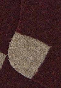 Nahaufnahme eines burgunderfarbenen Strickstoffs mit einem beigen rechteckigen Patch, der weiche, strukturierte Wollfasern zeigt.