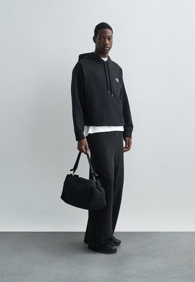 Hanorac negru cu un logo, suprapus peste un tricou alb. Asortat cu pantaloni negri largi. Ține în mână o geantă de mână neagră, structurată. Material texturat.