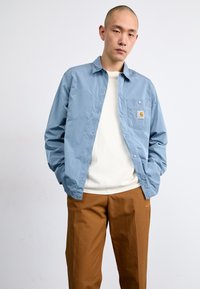 Veste boutonnière bleu clair avec une poche avant, portée sur une chemise crème, associée à un pantalon marron ; texture de tissu lisse et léger.