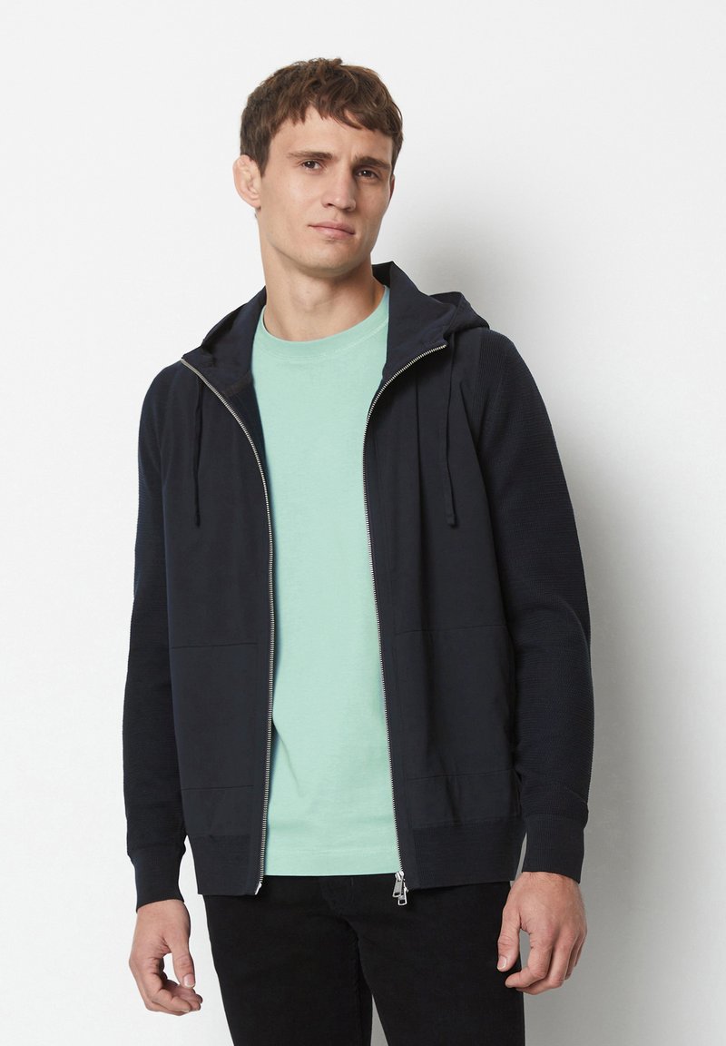 Dark navy zip-up hoodie with a front pocket, ribbed cuffs. Worn over a light mint t-shirt, showcasing a simple, casual design.