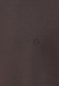 Camiseta de algodón marrón con una textura suave que presenta un pequeño logo circular bordado en un color contrastante.