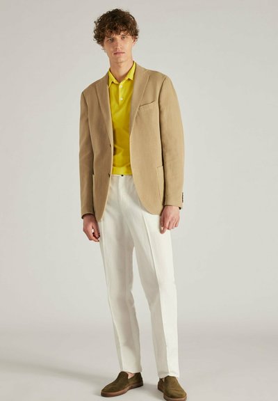 Beige blazer over een gele polo, gecombineerd met witte broeken en groene instappers. De stof heeft een gladde textuur met zachte lijnen.