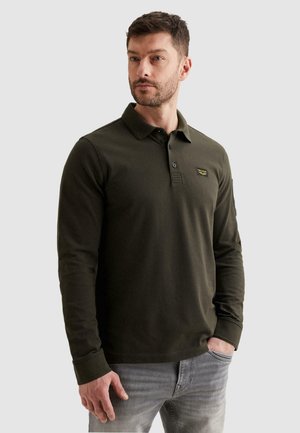 Muškarac odjeven u tamnozelenu polo majicu dugih rukava s ovratnikom i svijetlosive traperice, stoji s jednom rukom u džepu nasuprot jednostavnoj pozadini.