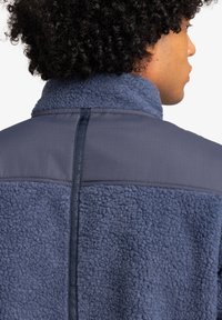 Fleece jacket in dark navy featuring a textured body, smooth accent panel, and a high collar with vertical brand detailing on the back.