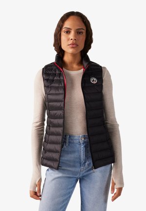 Mujer con un chaleco negro acolchado sin mangas sobre una camiseta beige de manga larga y vaqueros azul claro, de pie frente a un fondo sencillo.