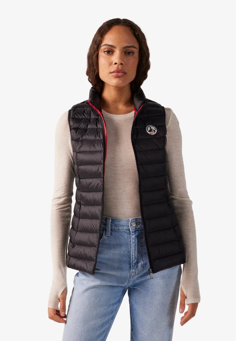 Mujer con un chaleco negro acolchado sin mangas sobre una camiseta beige de manga larga y vaqueros azul claro, de pie frente a un fondo sencillo.