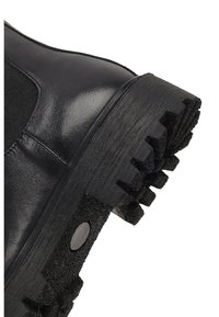 Bottine en cuir noir avec panneau latéral élastique, semelle en caoutchouc texturée avec un profond relief, et un accent métallique circulaire sur le talon.