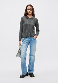 Cardigan gris de punto con cuello y botones delanteros, combinado con vaqueros sueltos azul claro. La modelo sostiene un pequeño bolso gris. Zapatos negros gruesos.