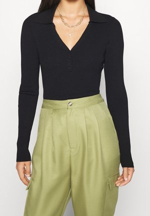 Femme portant un haut noir à manches longues côtelé avec un col en V boutonné et deux colliers dorés superposés, assorti à un pantalon taille haute vert olive.