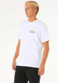 T-shirt bianca in cotone con una piccola grafica in testo dorato sul petto. Maniche corte con collo tondo, vestibilità regolare.