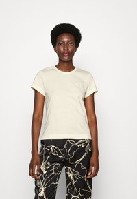 Camiseta de algodón de manga corta, color beige claro, con Escote redondo y un pequeño detalle gráfico en el lado izquierdo del pecho. Combinada con pantalones negros de un patrón.