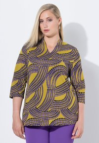 Driekwartmouwen blouse in mosterdgeel met golvende paarse en zwarte stippenpatronen. V-hals ontwerp met een soepele textuur.